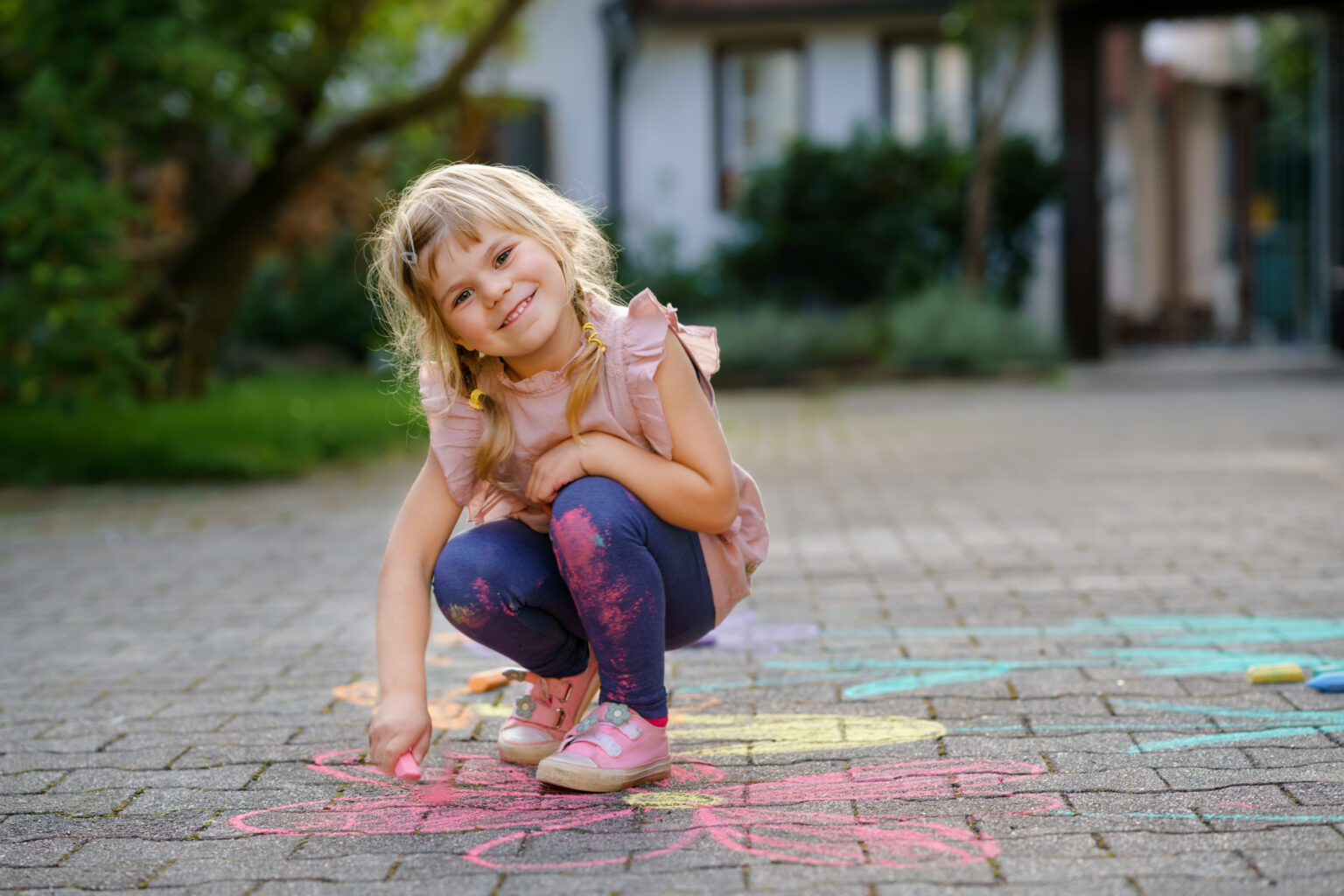 Little preschool girl painting with colorful chalks flowers on ground on backyard. Positive happy toddler child drawing and creating pictures on asphalt. Creative outdoors children activity in summer